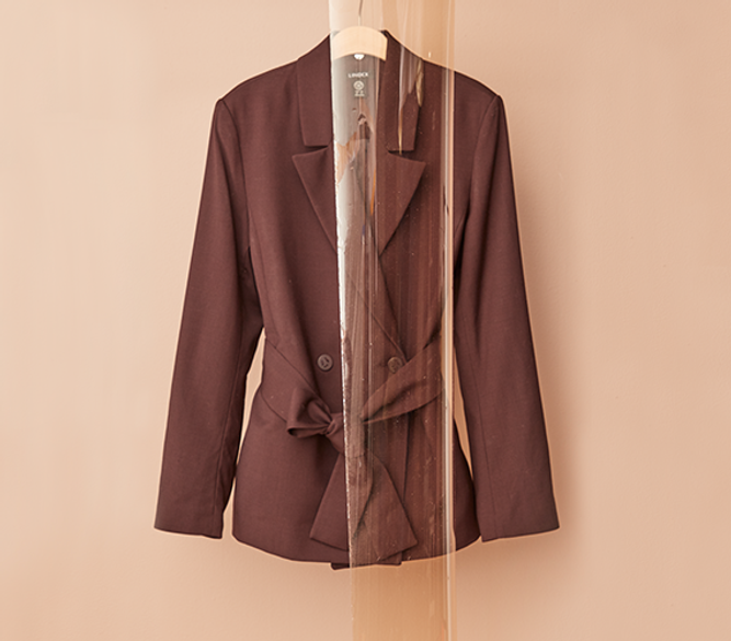 A brown blazer with a tie belt hangs on a wooden hanger against a beige background with a see through piece infront of it.
