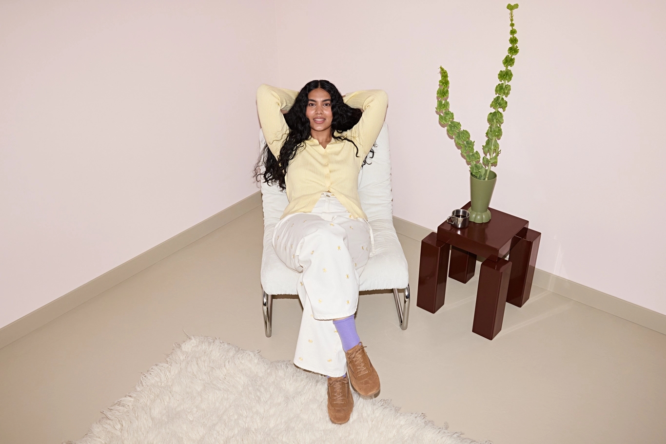 Person sitting on a pink chair, wearing a pink shirt and track pants with white stripes. They are resting their chin on their hand, with a neutral expression, in a room with blue curtains.